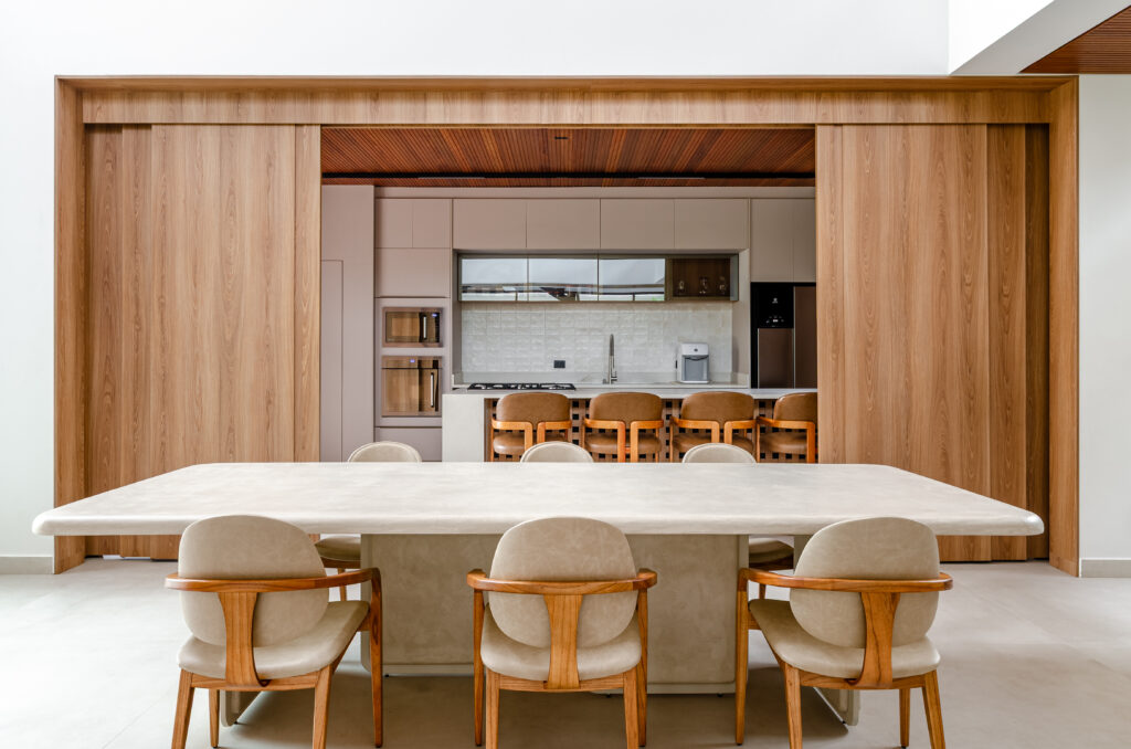 Sala de jantar com painel de portas de correr em madeira aberto, revelando a cozinha – Casa Sintonia