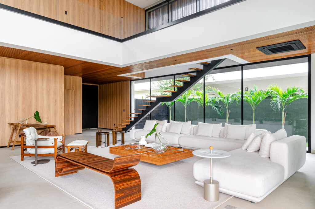 Living de pé-direito duplo com escada de degraus de madeira soltos e jardim tropical ao fundo – Casa Sintonia