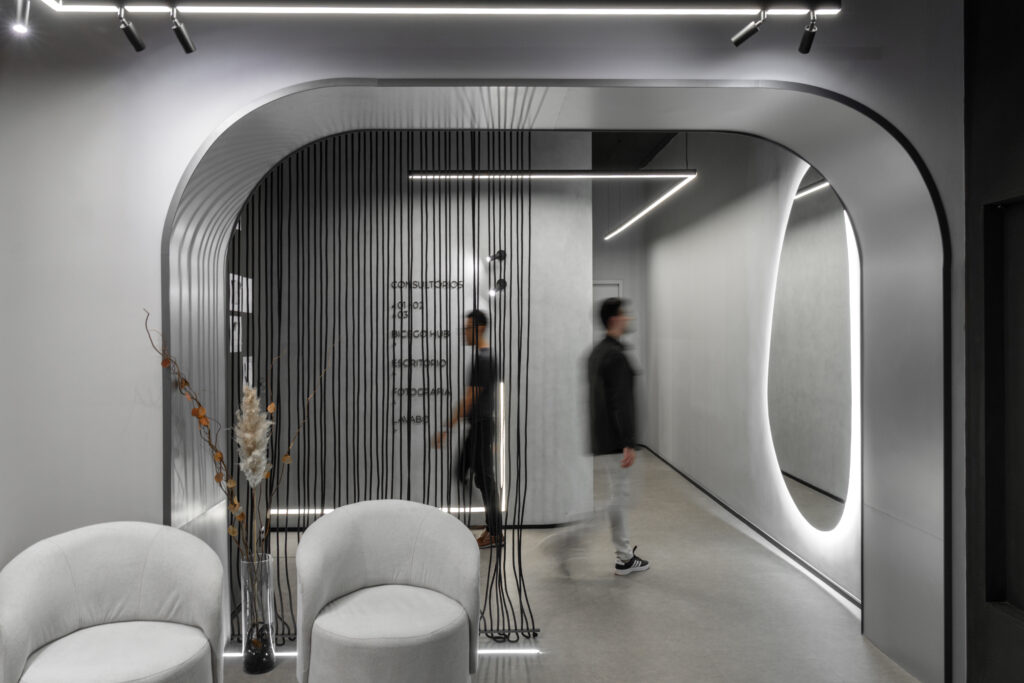 Corredor de entrada curvo com fitas pretas suspensas, poltronas brancas e espelho orgânico iluminado – Bicego Clinic